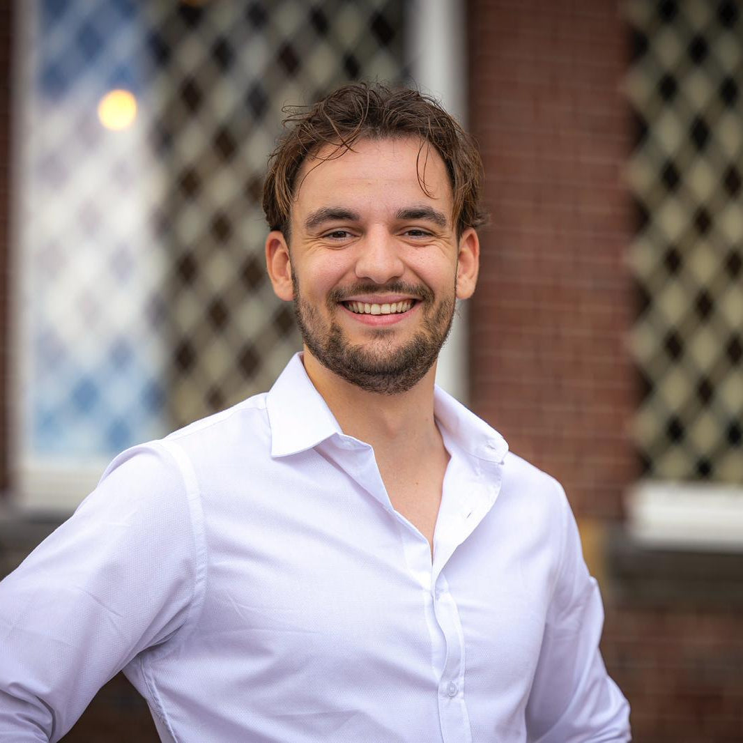 Lucas wearing a white shirt standing in front of a brick wall with decorative windows.
