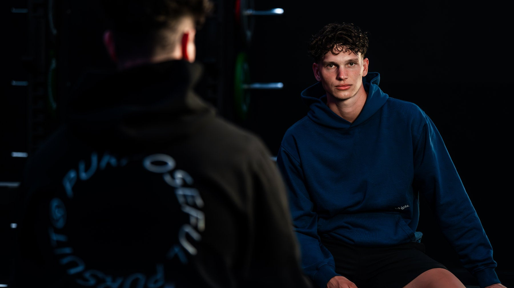 Banner Models sitting forwards and backwards and have on the orangic cotton Prelude Hoodie Black and Indigo Blue Origin. in a dark setting.