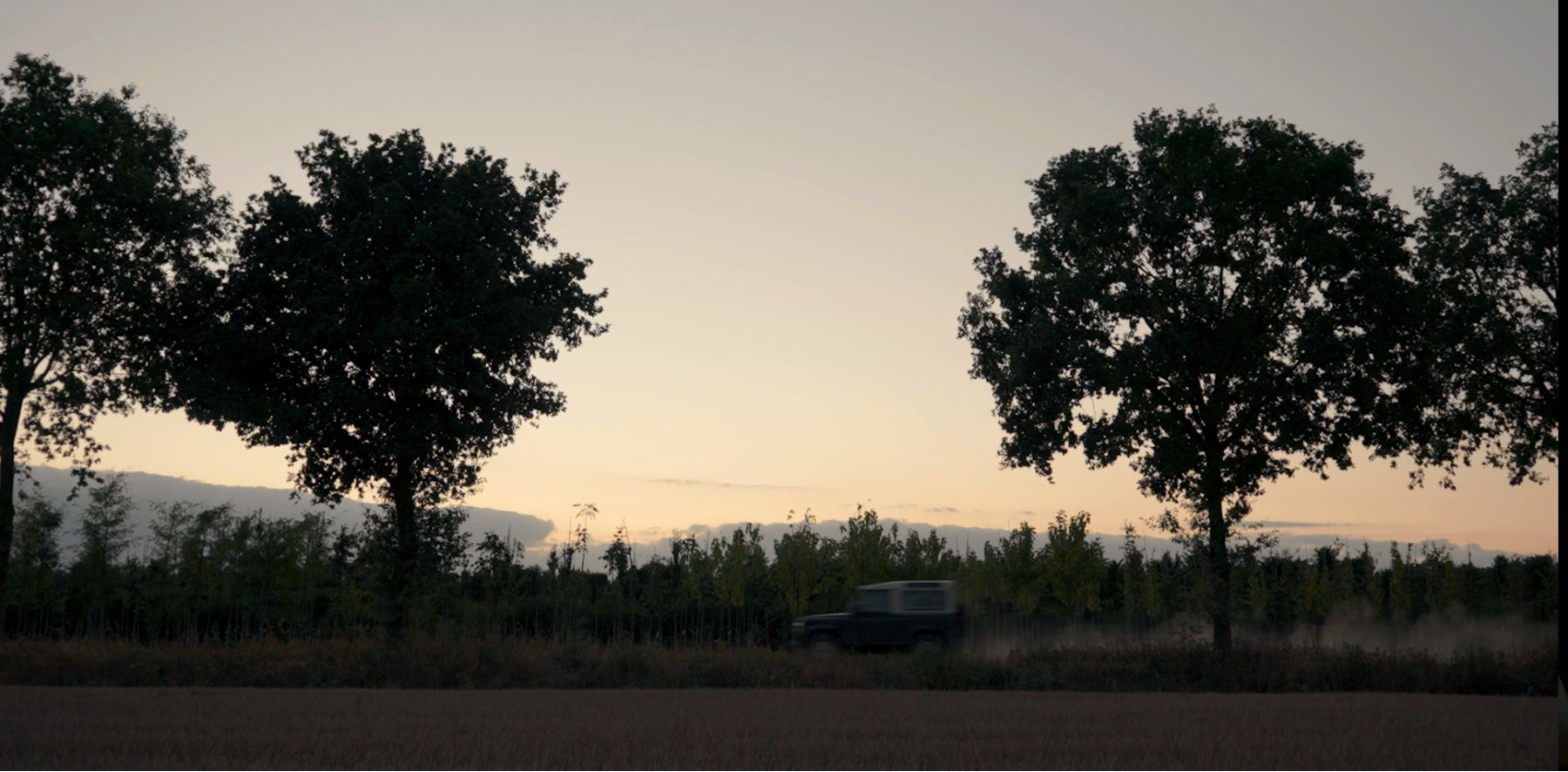 Land Rover Defender drives along a dirt track through the trees at sunset during a campaign scene from Origin. 