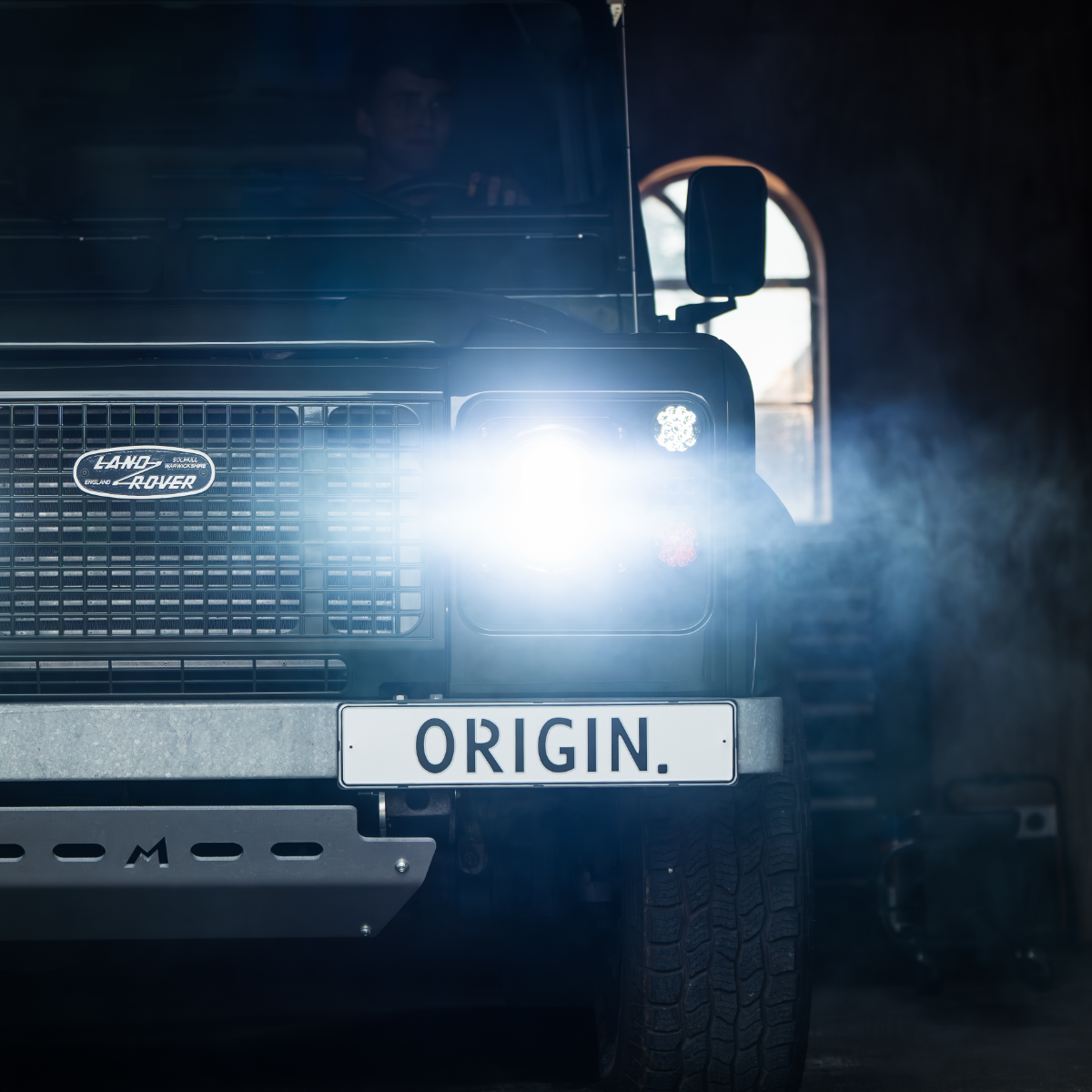 Front view of a Land Rover Defender vehicle with 'Origin.' license plate in a dimly lit garage.