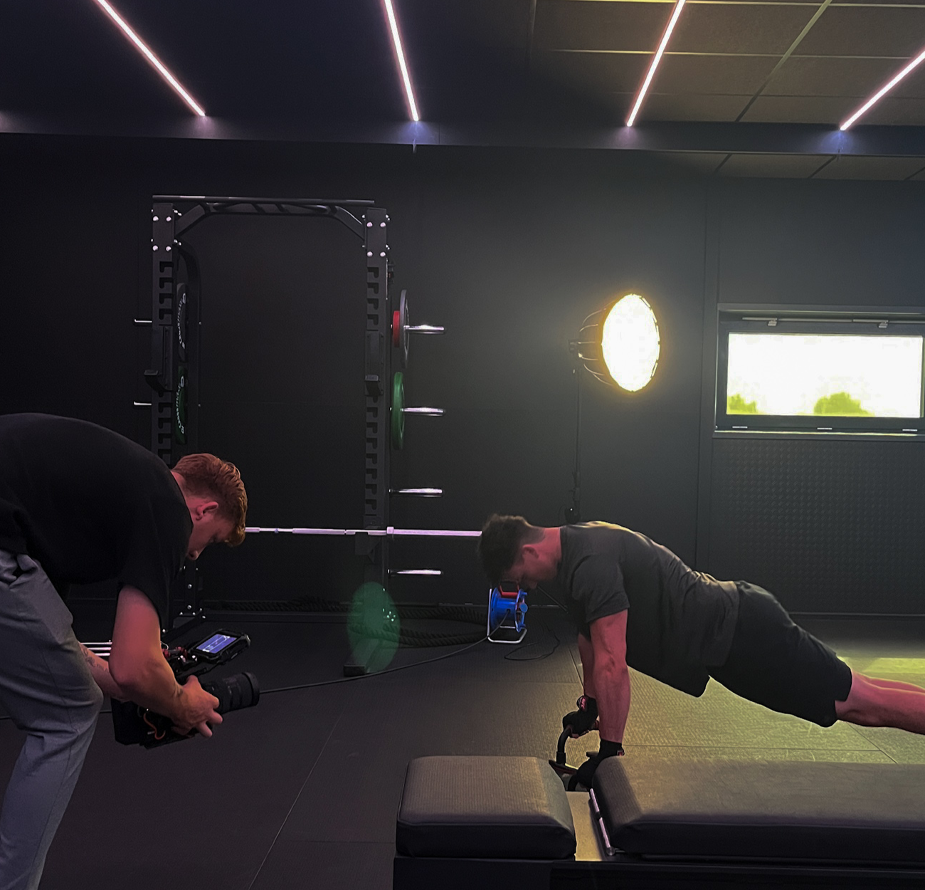 Origin. crew in a dark gym with shoot equipments, lights and model doing push-ups during he is filming by videographer.