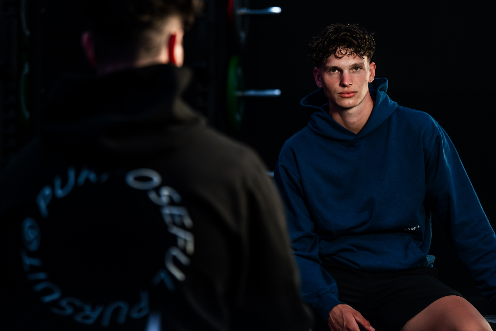 Models sitting forwards and backwards and have on the organic cotton Prelude Hoodie Black and Indigo Blue from Origin. in a dark setting.