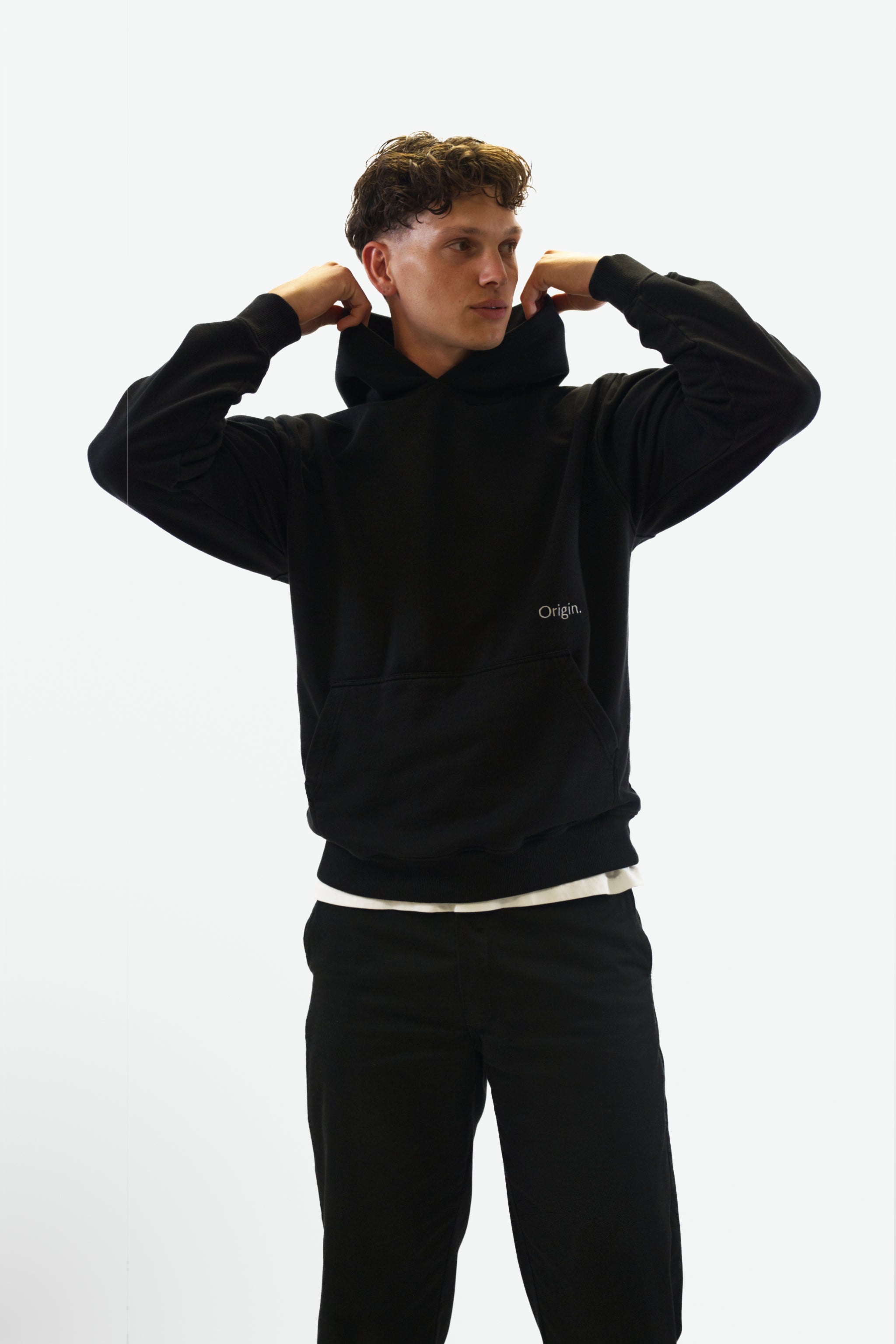 Front view grab the hood of model wearing the Prelude Hoodie in black from Origin. made from organic cotton in a clean studio setting.