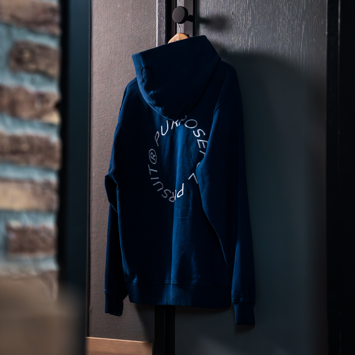 Prelude Hoodie Indigo Blue Origin. organic cotton hooded sweater with back embroidery, hanging on wooden hanger on a door.