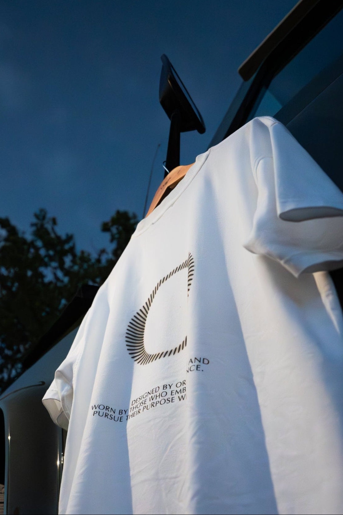 The Slash Tee White Origin. organic cotton t-shirt with graphic print, hanging against a Land Rover Defender door with a blue sky in the background.