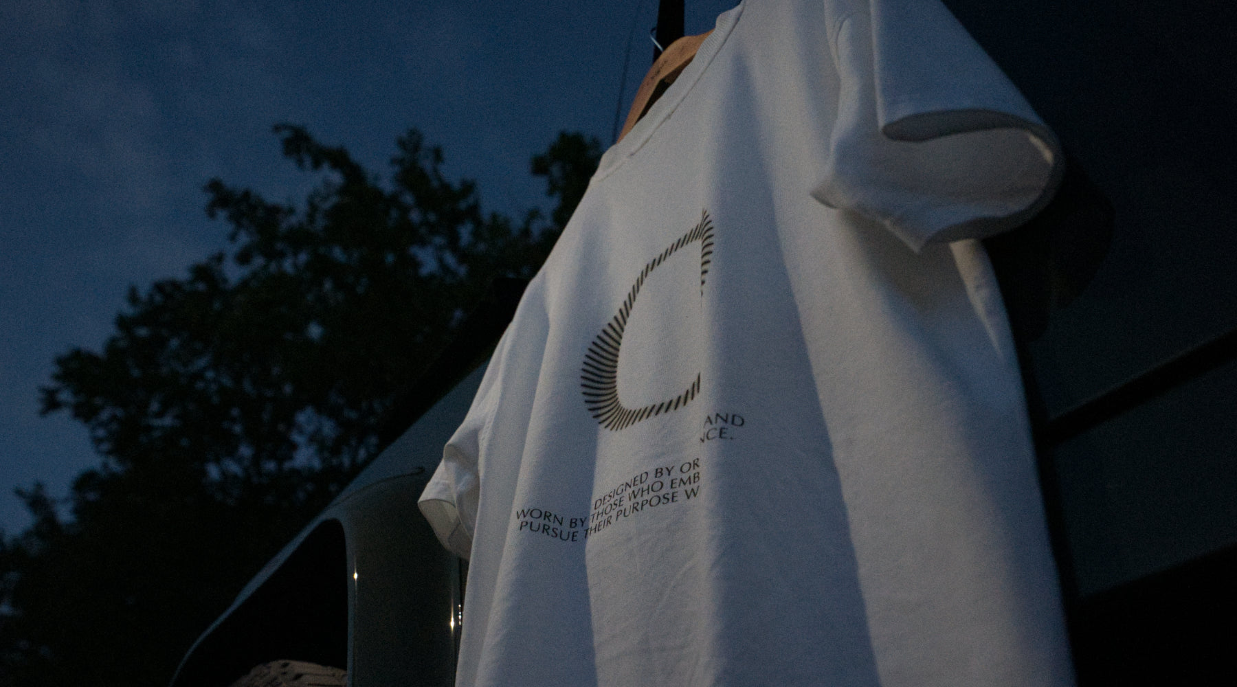 The Slash Tee White Origin. organic cotton t-shirt with graphic print, hanging against a Land Rover Defender door with a blue sky in the background edited in foggy view.