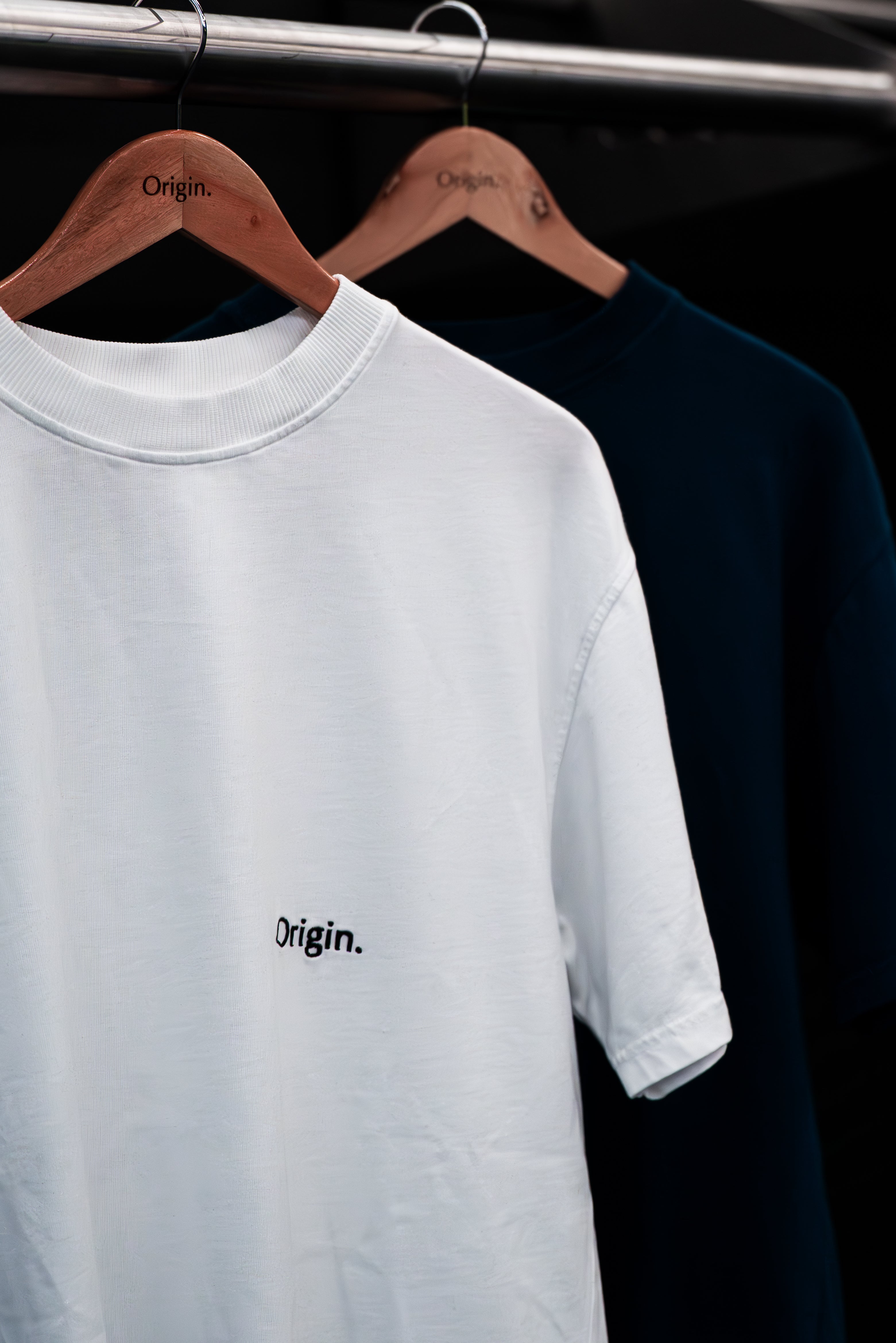 White and Indigo Blue Tee  with 'Origin.' logo on a wooden hanger against a dark background.