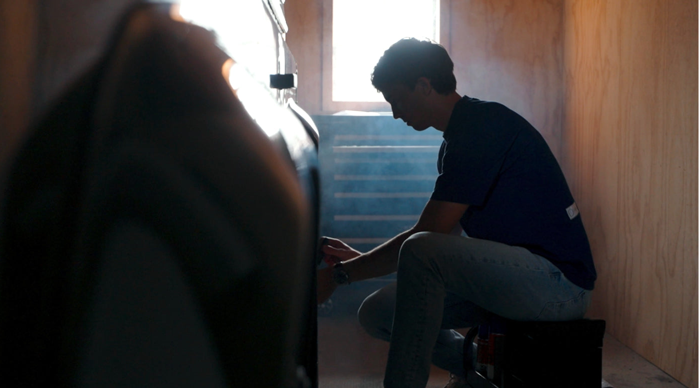 A person sits on a box, working with a tool beside a Land Rover Defender in a dimly lit room, wearing the Foundation Tee in Indigo Blue from Origin.