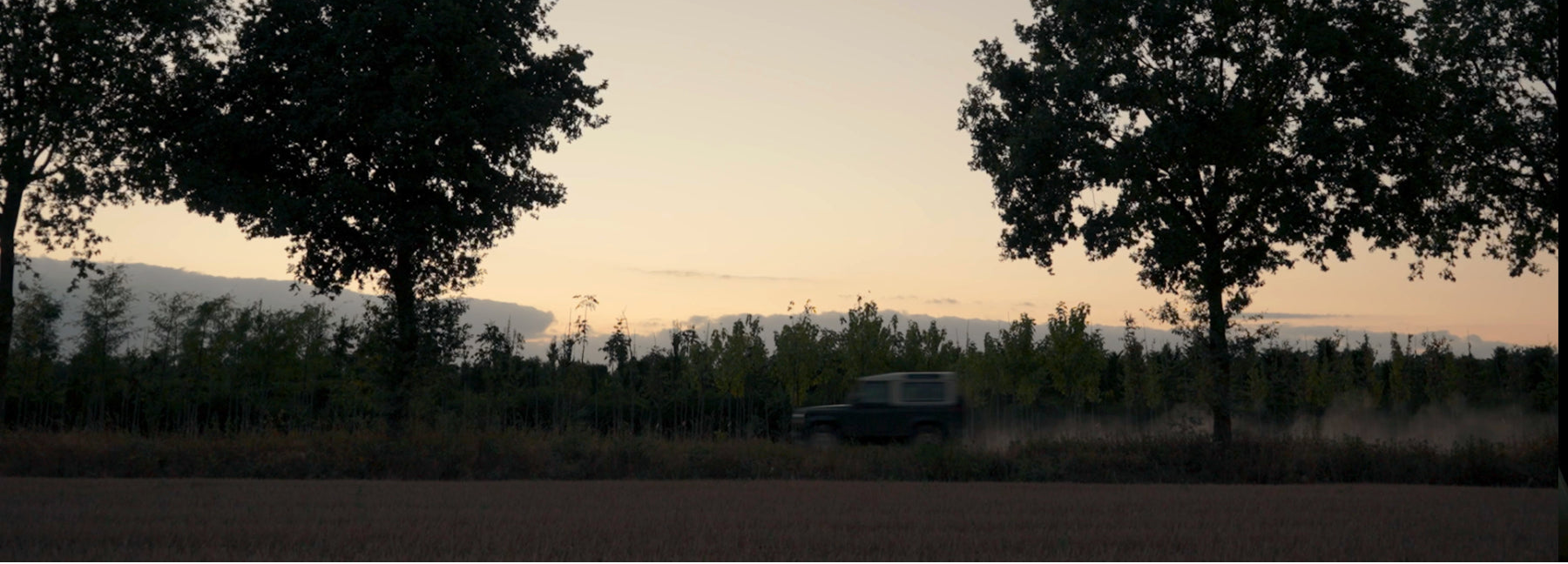 Silhouette of trees against a sunset sky with a Land Rover Defender in the distance.