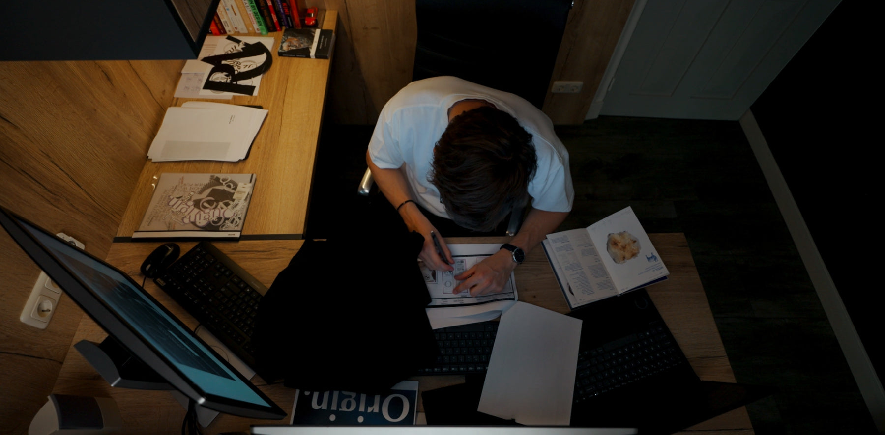 Person sitting at a desk with a computer and various items on a wooden floor. The person wearing a Origin. Slash Tee White.